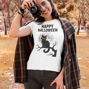 May include: White t-shirt with the text "HAPPY HALLOWEEN" above a black cat perched on a tree branch in front of a full moon. The cat has green eyes. The shirt is worn by a person.