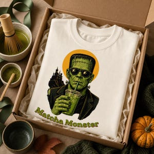 Może przedstawiać: Kremowy t-shirt z grafiką zielonego potwora w czarnych okularach przeciwsłonecznych, pijącego z przezroczystego kubka przez słomkę. Za potworem znajduje się ż&oacute;łte koło i sylwetka ciemnego zamku. Poniżej nadrukowany napis "Matcha Monster".