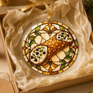 May include: A round, clear ornament featuring a stained-glass style illustration of a cannoli filled with cream, chocolate chips, and pistachios. The design is framed by amber, green, and cream-colored geometric patterns and hangs by a gold ribbon.
