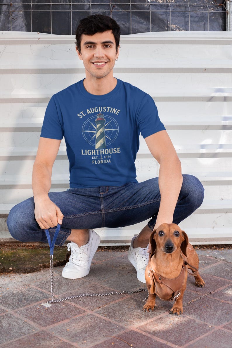 May include: A blue t-shirt with a graphic of a lighthouse, compass, and the words 'St. Augustine Lighthouse Est. 1874 Florida.' The person is wearing blue jeans and white sneakers, squatting next to a brown dachshund wearing a harness and leash.