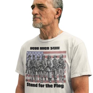 May include: A light-colored t-shirt featuring a graphic of seven skeleton soldiers in various historical military uniforms standing in front of an American flag. The text "Dead Men Still" appears above the graphic, and "Stand for the Flag" is below.