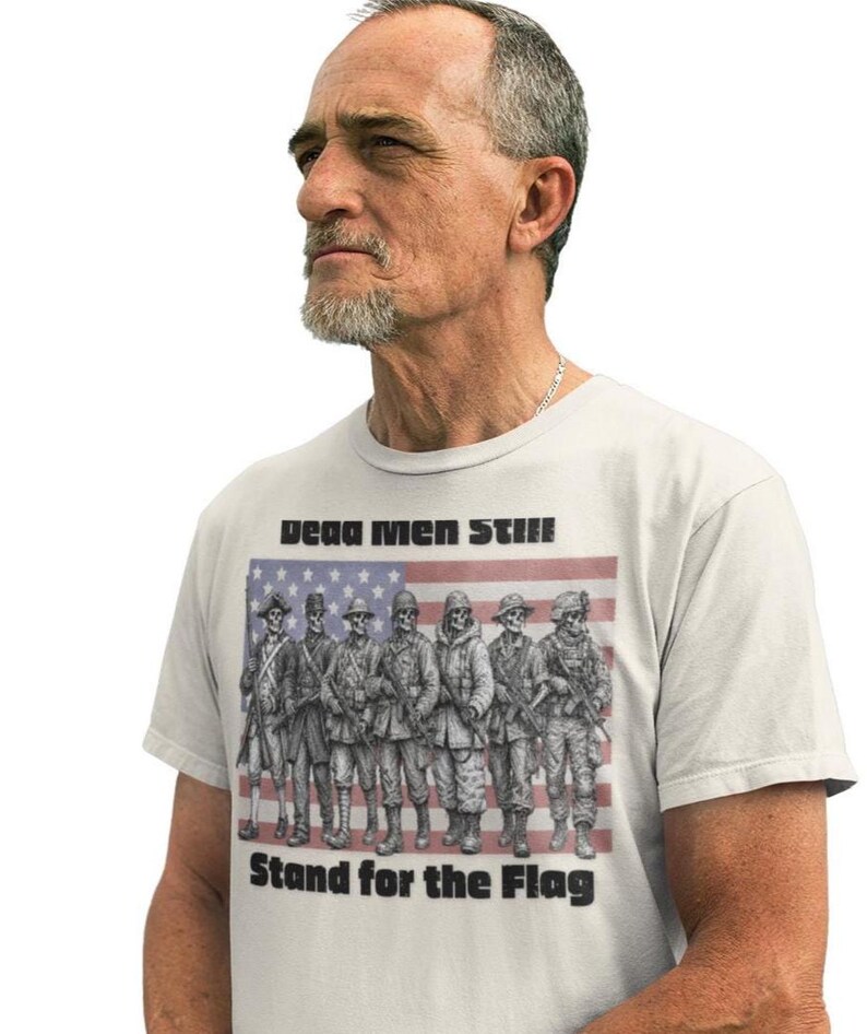May include: A light-colored t-shirt featuring a graphic of seven skeleton soldiers in various historical military uniforms standing in front of an American flag. The text "Dead Men Still" appears above the graphic, and "Stand for the Flag" is below.