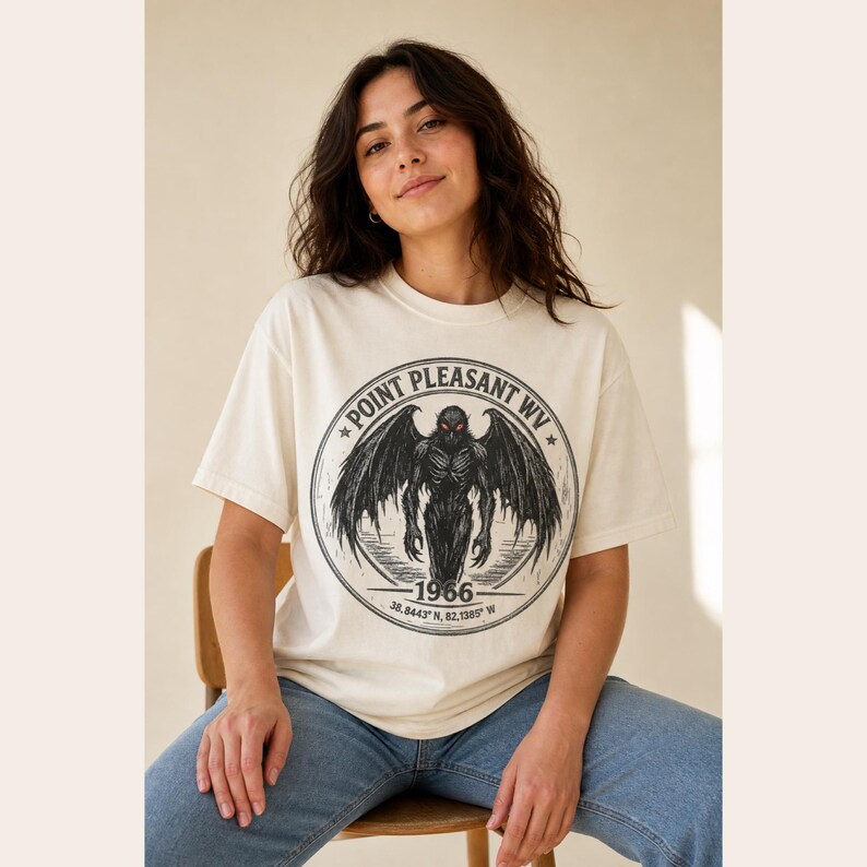 May include: A cream-colored short-sleeve t-shirt featuring a circular black graphic of a winged creature with red eyes. The text reads Point Pleasant WV, 1966, and the coordinates 38.8443 N, 82.1385 W, printed in a distressed black font.