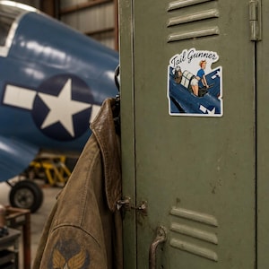 May include: A die-cut vinyl sticker featuring a pin-up style illustration of a person operating a machine gun in the cockpit of a blue military aircraft. The sticker includes the text Tail Gunner in a cursive script font at the top.