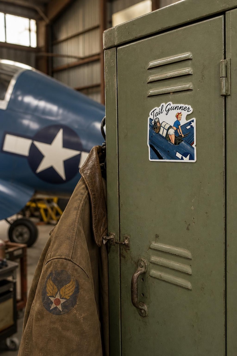 May include: A die-cut vinyl sticker featuring a pin-up style illustration of a person operating a machine gun in the cockpit of a blue military aircraft. The sticker includes the text Tail Gunner in a cursive script font at the top.