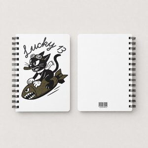 May include: A white spiral-bound notebook featuring a black and white illustration of a cat wearing goggles and a scarf while riding a bomb. The text Lucky 13 is printed above the cat. The back cover is plain white with a small barcode at the bottom.