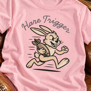 Puede incluir: Una camiseta de manga corta de color rosa claro con un gr&aacute;fico de un conejo de dibujos animados de color beige corriendo mientras sostiene una bomba marr&oacute;n. Sobre el conejo, el texto Hare Trigger est&aacute; impreso en una fuente cursiva gris oscuro.