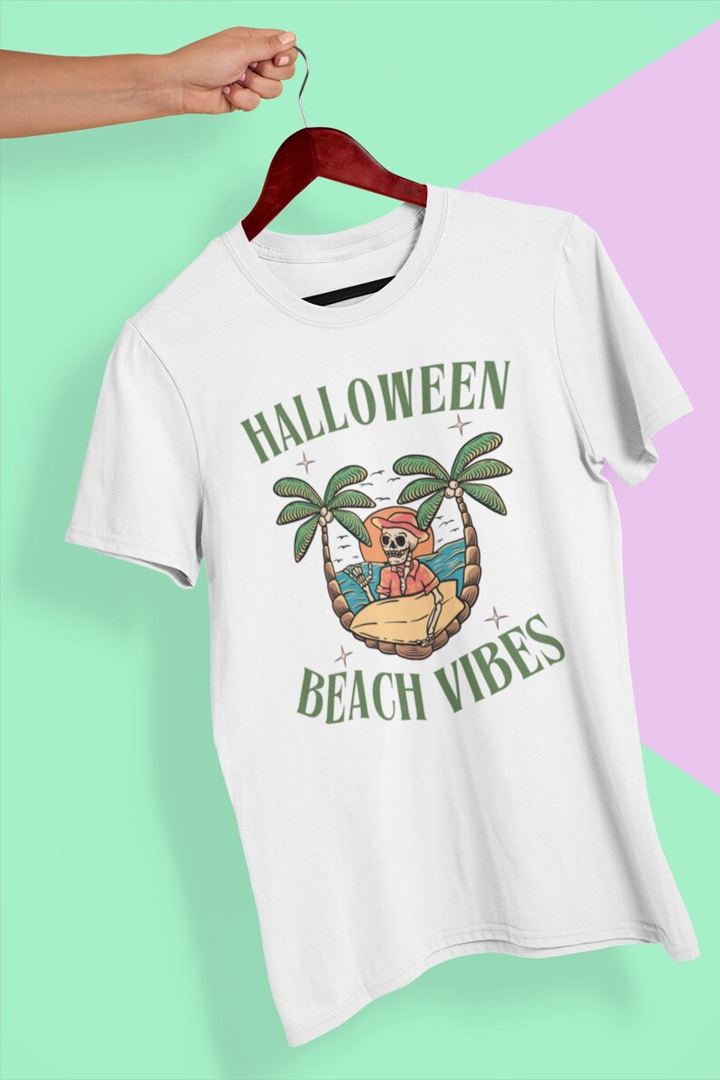 May include: White t-shirt with a Halloween graphic. The design shows a skeleton on a beach with palm trees, the sun, and the words "HALLOWEEN BEACH VIBES" in green.