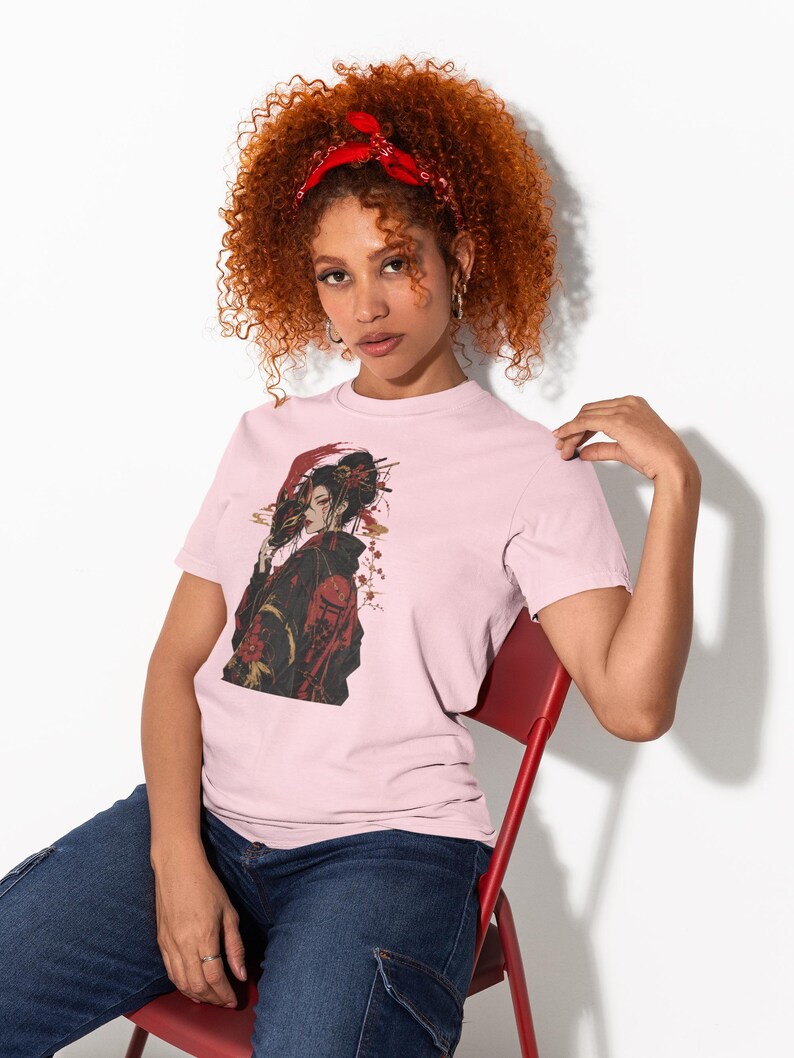 May include: A light pink short-sleeved t-shirt featuring a graphic print of a person with dark hair styled in an updo, wearing a black and red patterned garment, holding a red mask, with red floral accents and a small red torii gate design.