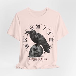 May include: Light pink t-shirt featuring a black illustration of a raven perched atop a skull, set against a clock face with Roman numerals. The words "Memento Mori" and the Roman numeral "MMXXV" are printed below the skull.