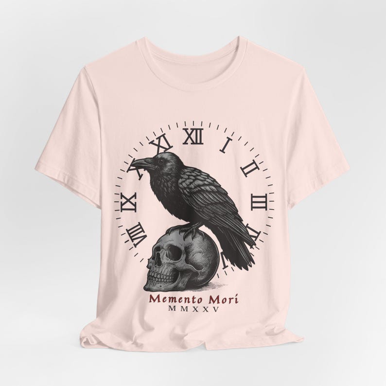 May include: Light pink t-shirt featuring a black illustration of a raven perched atop a skull, set against a clock face with Roman numerals. The words "Memento Mori" and the Roman numeral "MMXXV" are printed below the skull.