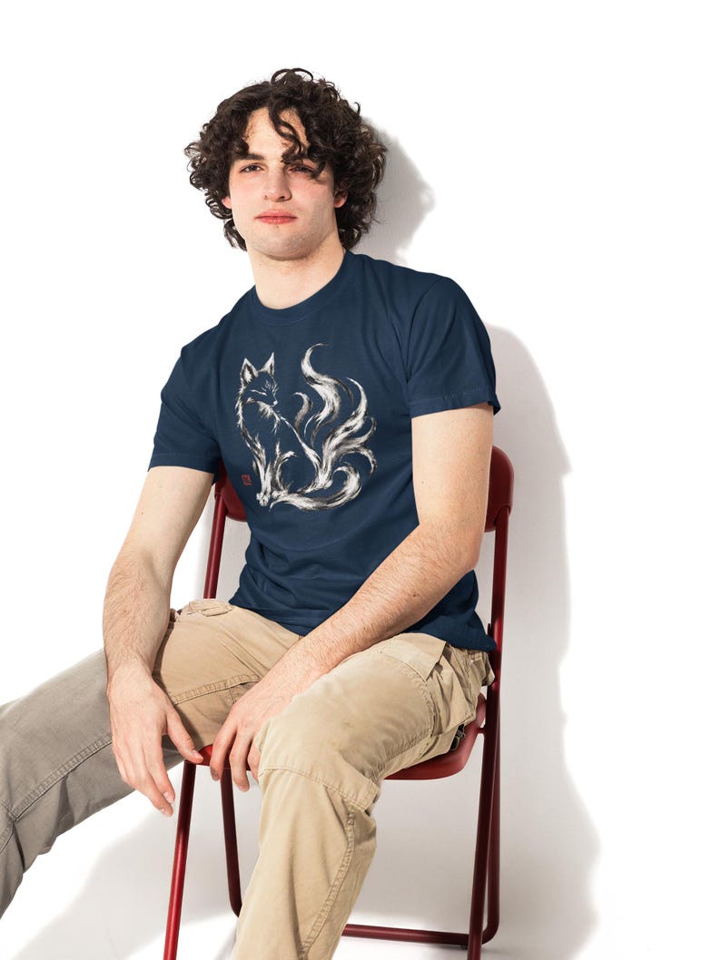 May include: A navy blue short-sleeved t-shirt featuring a white, brushstroke-style graphic of a multi-tailed fox sitting in profile. The design includes a small red square stamp detail near the base of the fox illustration on the chest.