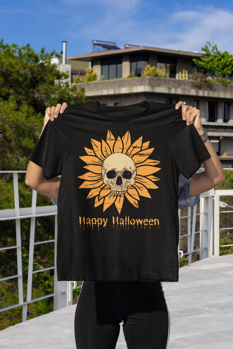 May include: Black t-shirt featuring a sunflower design with a skull in the center. The sunflower is orange, and the skull is off-white. The words "Happy Halloween" are printed below the design in orange.