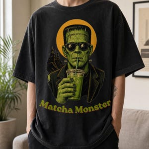 Może przedstawiać: Ciemnoszary t-shirt z kr&oacute;tkim rękawem z nadrukiem przedstawiającym zielonego potwora w okularach przeciwsłonecznych, pijącego nap&oacute;j przez słomkę. Za postacią znajduje się ż&oacute;łte koło i sylwetka zamku. Poniżej znajduje się napis Matcha Monster.