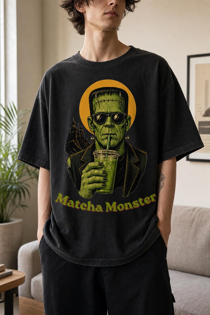 Może przedstawiać: Ciemnoszary t-shirt z kr&oacute;tkim rękawem z nadrukiem przedstawiającym zielonego potwora w okularach przeciwsłonecznych, pijącego nap&oacute;j przez słomkę. Za postacią znajduje się ż&oacute;łte koło i sylwetka zamku. Poniżej znajduje się napis Matcha Monster.