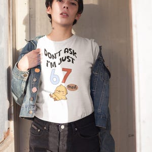 Puede incluir: Camiseta blanca con el texto "DON'T ASK I'M JUST 67 meh" y una ilustraci&oacute;n de pollito de dibujos animados. La camiseta se lleva con una chaqueta vaquera y pantalones cortos negros.