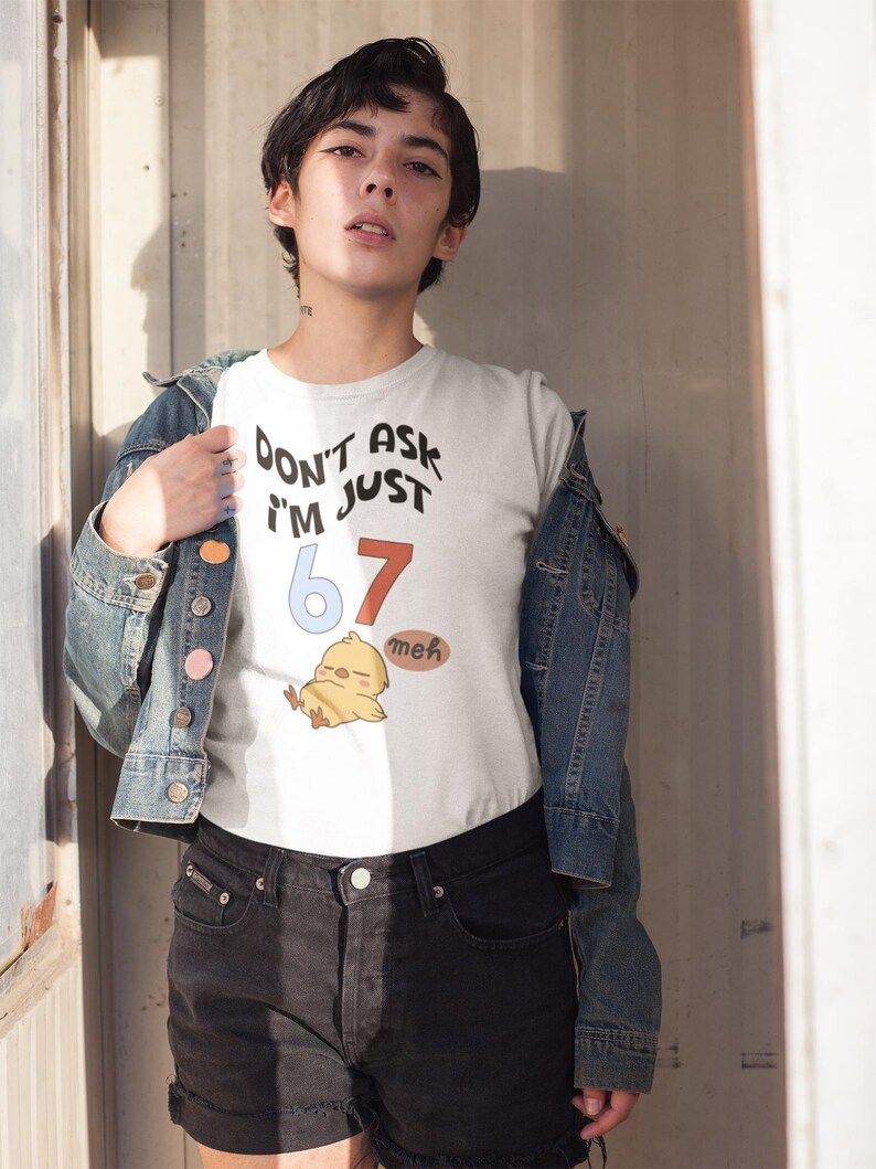 Puede incluir: Camiseta blanca con el texto "DON'T ASK I'M JUST 67 meh" y una ilustraci&oacute;n de pollito de dibujos animados. La camiseta se lleva con una chaqueta vaquera y pantalones cortos negros.