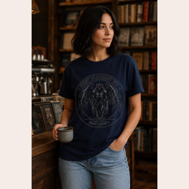 May include: A navy blue short-sleeve t-shirt featuring a circular graphic of a winged creature with red eyes. The text reads Point Pleasant WV and 1966. The design is rendered in shades of gray and white, centered on the chest of the garment.