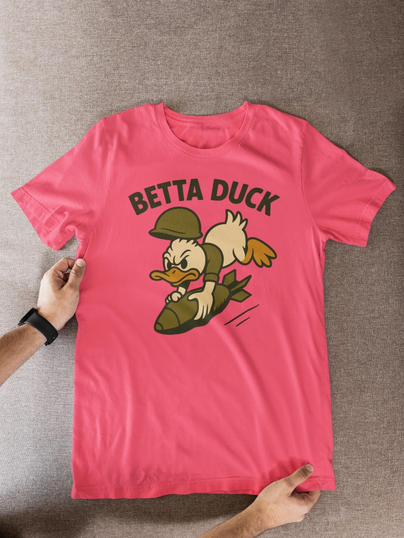 May include: Pink t-shirt with a cartoon duck riding a bomb, and the words "BETTA DUCK" above. The duck wears a military helmet and uniform. The shirt is held up by two hands, with a black watch on one wrist.