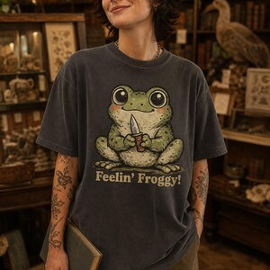 Może przedstawiać: Ciemnoszary t-shirt z kr&oacute;tkim rękawem z grafiką przedstawiającą zieloną żabę trzymającą mały n&oacute;ż. Pod żabą znajduje się napis "Feelin' Froggy!" w jasnobeżowym kolorze. Koszulka posiada okrągły dekolt i luźny kr&oacute;j.