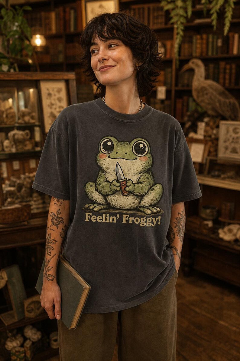Może przedstawiać: Ciemnoszary t-shirt z kr&oacute;tkim rękawem z grafiką przedstawiającą zieloną żabę trzymającą mały n&oacute;ż. Pod żabą znajduje się napis "Feelin' Froggy!" w jasnobeżowym kolorze. Koszulka posiada okrągły dekolt i luźny kr&oacute;j.