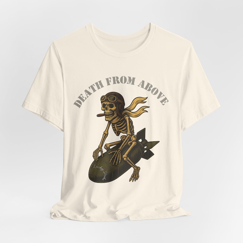 May include: Cream-colored t-shirt featuring a skeleton riding a bomb, wearing a pilot's cap and scarf, and smoking a cigar. The text "DEATH FROM ABOVE" arches above the skeleton. The design is in shades of brown, tan, and black.