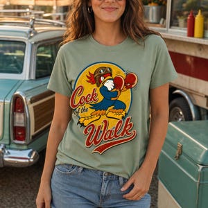 May include: A sage green t-shirt featuring a graphic of a cartoon rooster wearing aviator goggles and boxing gloves. The design is set within a yellow circle with a blue border and includes the red text Cock of the Walk in a script font.