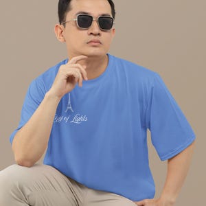 May include: A light blue t-shirt with the text "City of Lights" and an Eiffel Tower graphic. The shirt is short-sleeved and worn by a person wearing sunglasses and khaki pants.