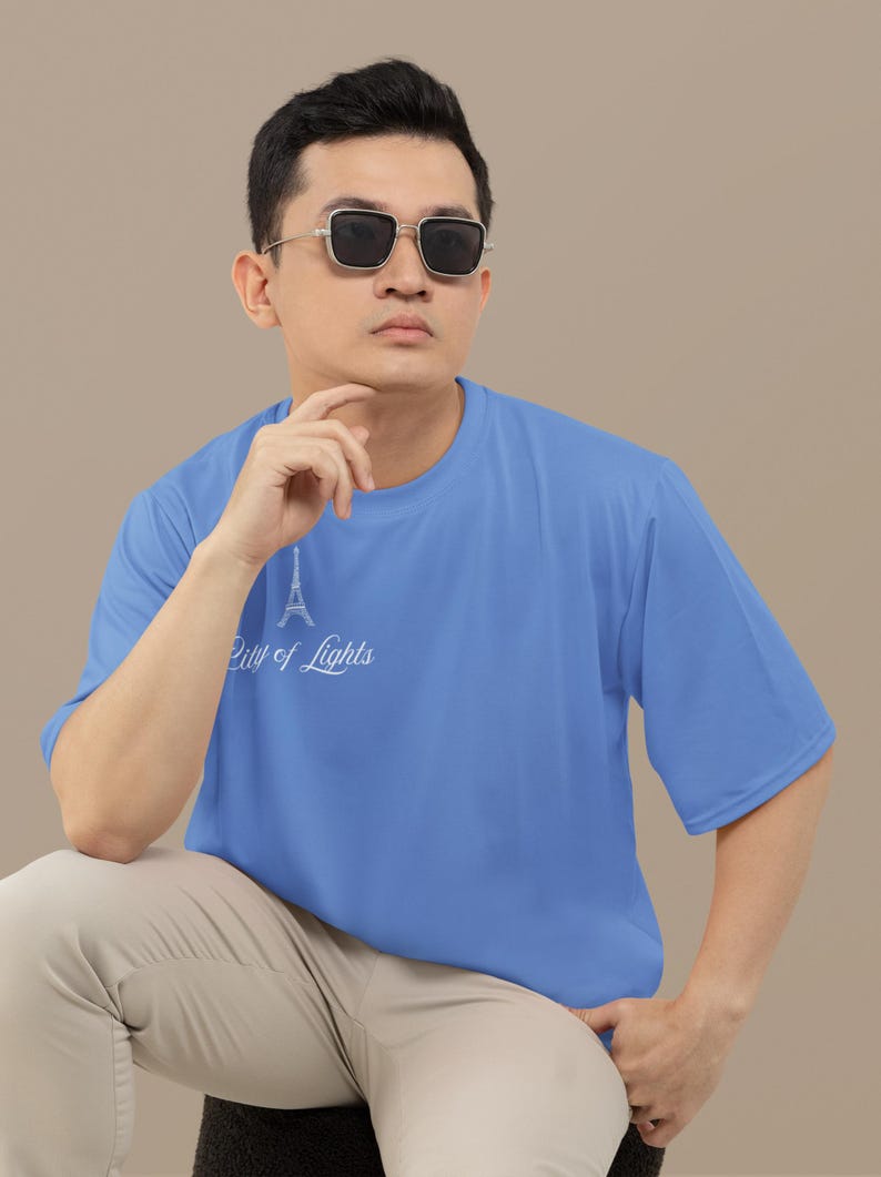 May include: A light blue t-shirt with the text "City of Lights" and an Eiffel Tower graphic. The shirt is short-sleeved and worn by a person wearing sunglasses and khaki pants.