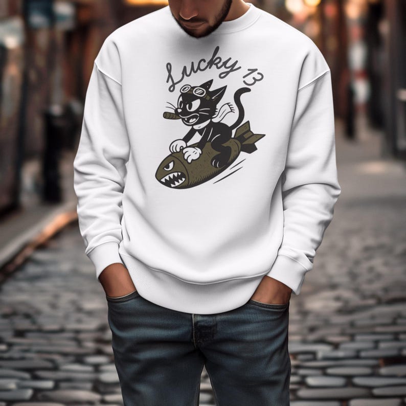 May include: White sweatshirt featuring a cartoon cat riding a bomb, with the text "Lucky 13" in a script font. The cat wears goggles and holds a cigar. The design is in black, olive green, and white.