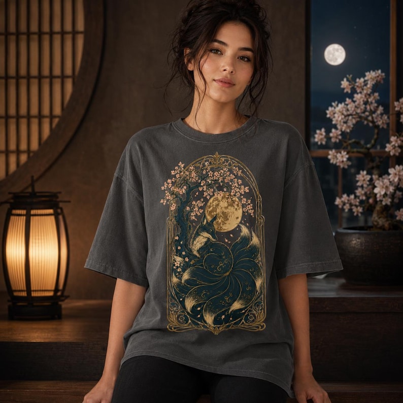 May include: A charcoal grey short-sleeve t-shirt featuring a central graphic print of a multi-tailed fox sitting beneath a flowering tree and a full moon, all enclosed within an ornate, gold-toned, arched frame with intricate decorative details.