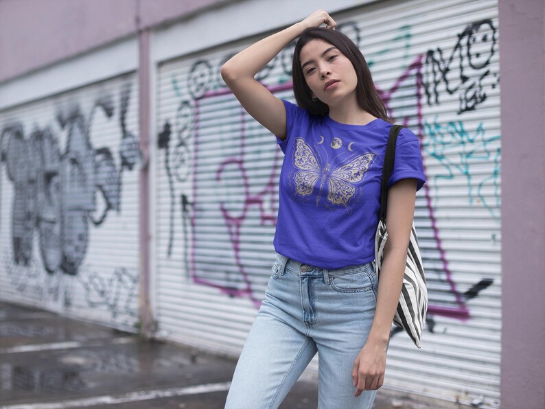 May include: A blue t-shirt featuring a butterfly design with moon phases. The butterfly is in gold, and the shirt is a solid blue color. The person is wearing light blue jeans and a black and white striped tote bag.