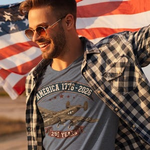 May include: A heather blue t-shirt featuring a graphic of a vintage military aircraft, the text AMERICA 1776-2026, and 250 YEARS. The shirt is worn under a black and white checkered button-down shirt, with a large American flag visible in the background.