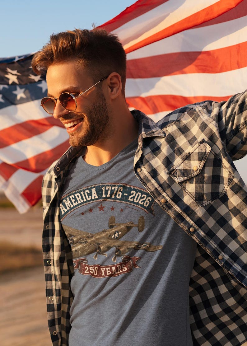 May include: A heather blue t-shirt featuring a graphic of a vintage military aircraft, the text AMERICA 1776-2026, and 250 YEARS. The shirt is worn under a black and white checkered button-down shirt, with a large American flag visible in the background.
