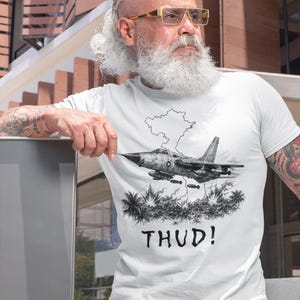 May include: White t-shirt featuring a black and white illustration of a jet fighter plane with the word "THUD!" below. The graphic includes a cloud and palm trees. The shirt is worn by a person.