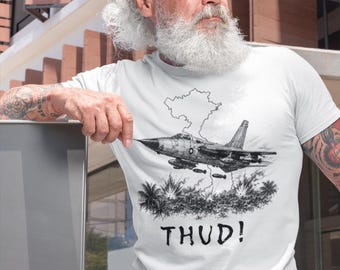 May include: White t-shirt featuring a black and white illustration of a jet fighter plane with the word "THUD!" below. The graphic includes a cloud and palm trees. The shirt is worn by a person.