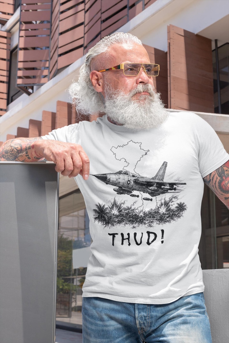 May include: White t-shirt featuring a black and white illustration of a jet fighter plane with the word "THUD!" below. The graphic includes a cloud and palm trees. The shirt is worn by a person.