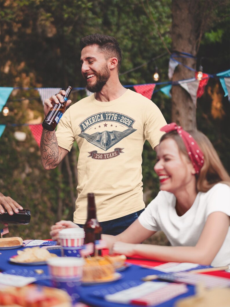 Puede incluir: Una camiseta de manga corta color crema con un estampado gr&aacute;fico de un avi&oacute;n bombardero furtivo sobre el texto AMERICA 1776-2026 y 250 YEARS, enmarcado por un dise&ntilde;o de pancarta con estrellas y una est&eacute;tica vintage desgastada.