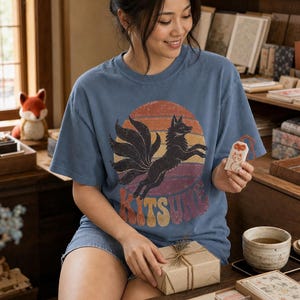 May include: A blue short-sleeved t-shirt featuring a graphic of a black nine-tailed fox leaping in front of a distressed sunset circle. The word KITSUNE is printed in large, retro-style block letters at the bottom of the graphic design.
