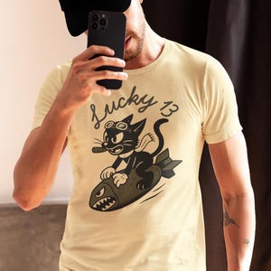May include: A cream-colored short-sleeve t-shirt featuring a graphic of a black cat wearing goggles and a scarf, riding a bomb with a shark-face design. The text "Lucky 13" is printed above the cat in a black script font on the front of the shirt.
