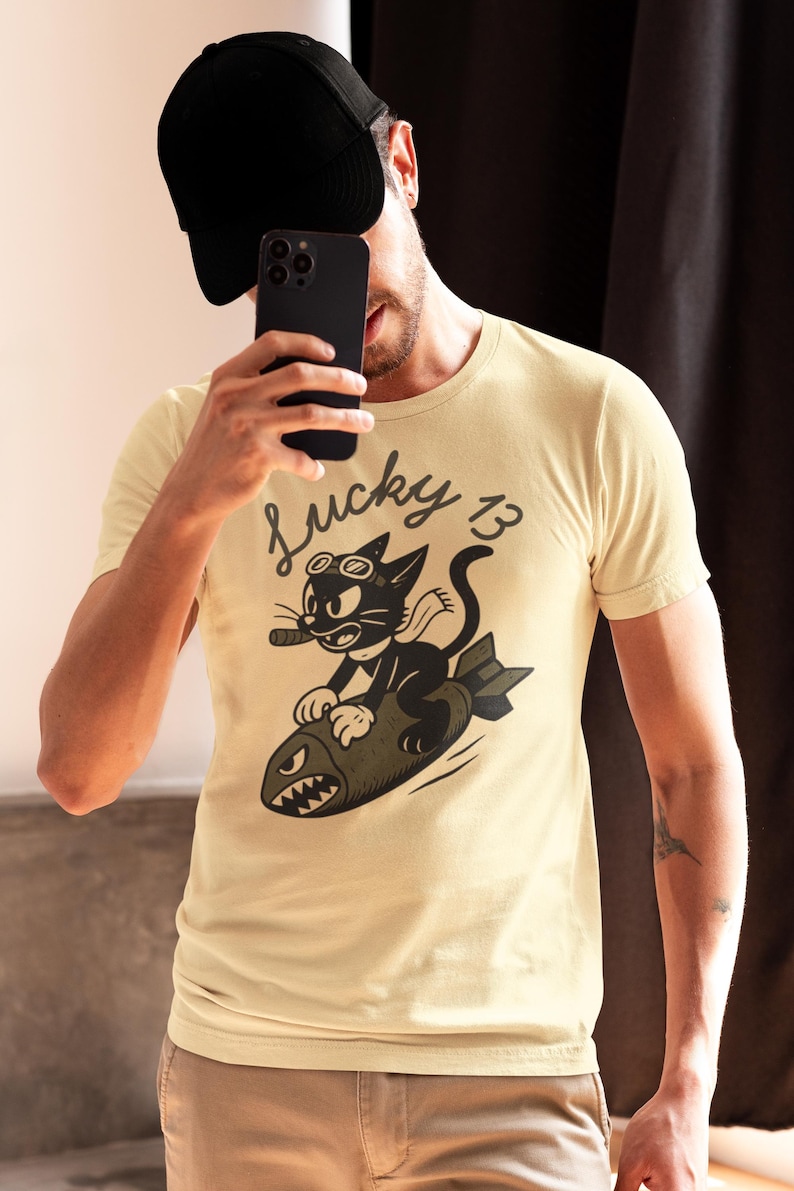 May include: A cream-colored short-sleeve t-shirt featuring a graphic of a black cat wearing goggles and a scarf, riding a bomb with a shark-face design. The text "Lucky 13" is printed above the cat in a black script font on the front of the shirt.