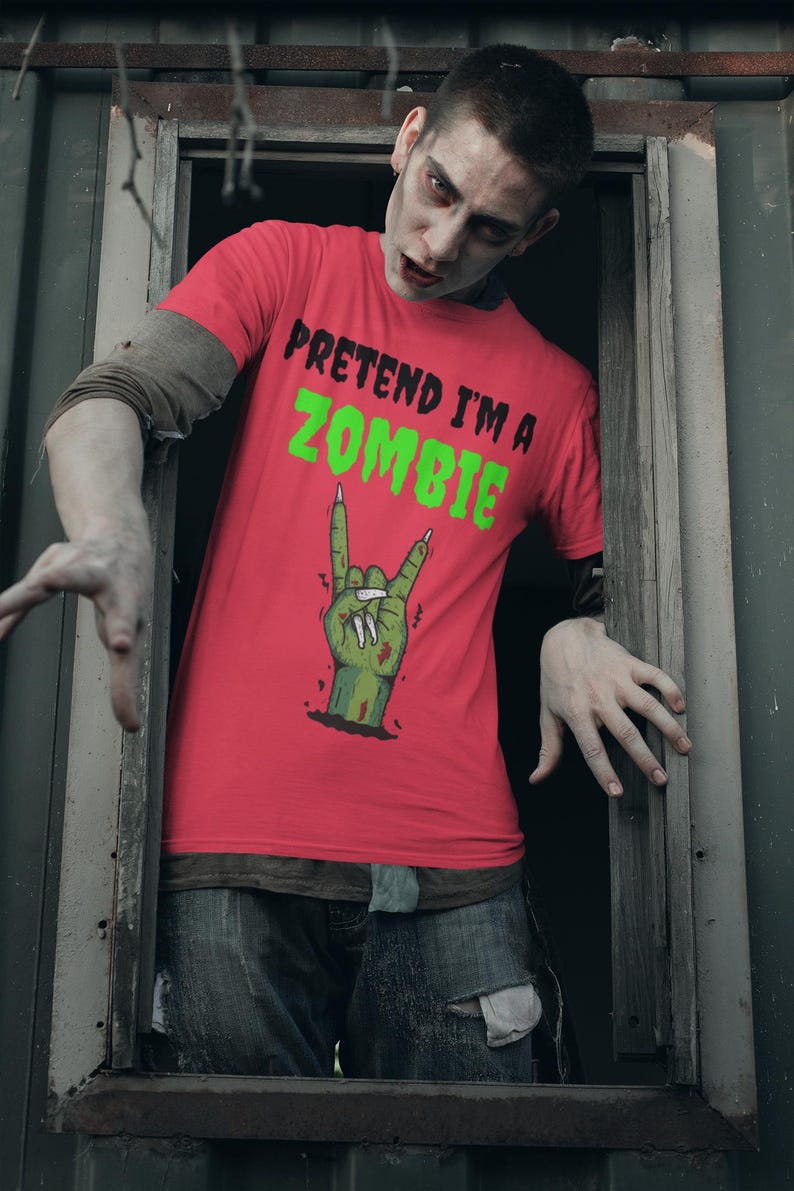 May include: A person in a red t-shirt with the words "PRETEND I'M A ZOMBIE" in black and green. The shirt features a green zombie hand making a rock and roll hand gesture. The person is wearing blue jeans and has zombie makeup.