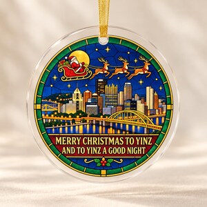 May include: A round, clear glass Christmas ornament featuring a stained-glass style illustration of the Pittsburgh skyline at night, Santa in a sleigh with reindeer, and the text MERRY CHRISTMAS TO YINZ AND TO YINZ A GOOD NIGHT.