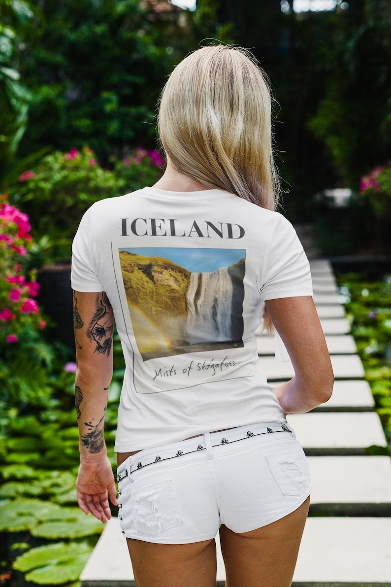 May include: A white t-shirt with a graphic of a waterfall and rainbow in Iceland. The word 'ICELAND' is printed above the image, and 'Mists of Skógafoss' is below. The person is wearing white shorts and standing on a stone pathway.
