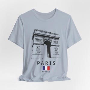 Puede incluir: Camiseta azul claro con un gr&aacute;fico en blanco y negro del Arco del Triunfo en Par&iacute;s. La palabra "PARIS" est&aacute; impresa debajo de la imagen, junto con la bandera francesa. Una camiseta informal para los amantes de los viajes.