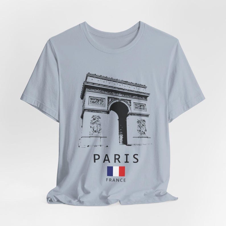 Puede incluir: Camiseta azul claro con un gr&aacute;fico en blanco y negro del Arco del Triunfo en Par&iacute;s. La palabra "PARIS" est&aacute; impresa debajo de la imagen, junto con la bandera francesa. Una camiseta informal para los amantes de los viajes.