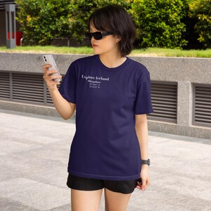May include: Navy blue t-shirt with the text "Explore Iceland" and coordinates. The shirt is short-sleeved and worn with black shorts. The person is wearing sunglasses and holding a smartphone.