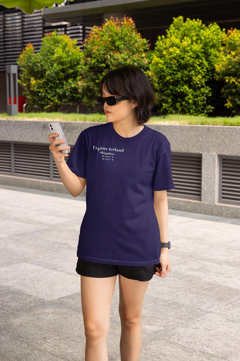 May include: Navy blue t-shirt with the text "Explore Iceland" and coordinates. The shirt is short-sleeved and worn with black shorts. The person is wearing sunglasses and holding a smartphone.