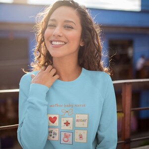 K&ouml;nnte beinhalten: Hellblaues Sweatshirt mit dem Text "mother baby nurse" und mehreren kleinen quadratischen Illustrationen. Die Illustrationen umfassen ein rotes Herz, ein Kreuz, Babyf&uuml;&szlig;e, einen Teddyb&auml;r und eine Babyflasche.