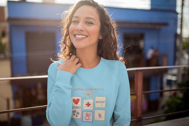 K&ouml;nnte beinhalten: Hellblaues Sweatshirt mit dem Text "mother baby nurse" und mehreren kleinen quadratischen Illustrationen. Die Illustrationen umfassen ein rotes Herz, ein Kreuz, Babyf&uuml;&szlig;e, einen Teddyb&auml;r und eine Babyflasche.